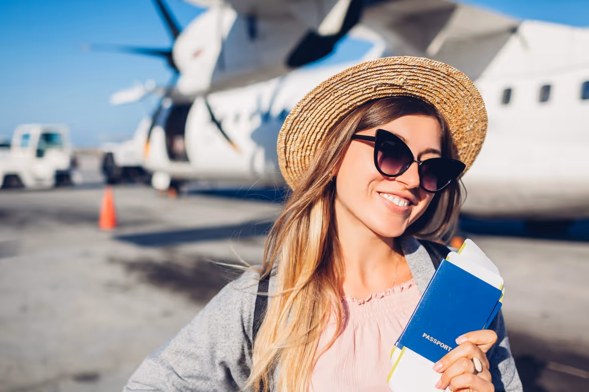 Jeune voyageuse souriante avec un passeport et un billet d’avion à la main devant un avion sur le tarmac.