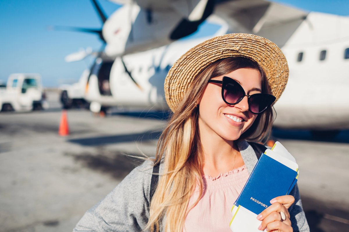 Jeune voyageuse souriante avec un passeport et un billet d’avion à la main devant un avion sur le tarmac.