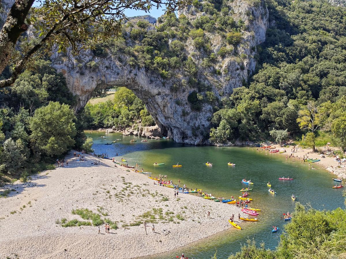 Vue du Pont d’Arc en Ardèche avec de nombreux canoës colorés sur la rivière et des vacanciers profitant de la plage.
