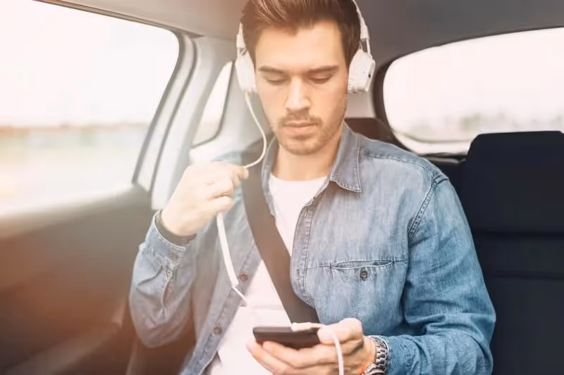 Un homme assis à l’arrière d’une voiture écoute de la musique avec un casque blanc, regardant son téléphone.