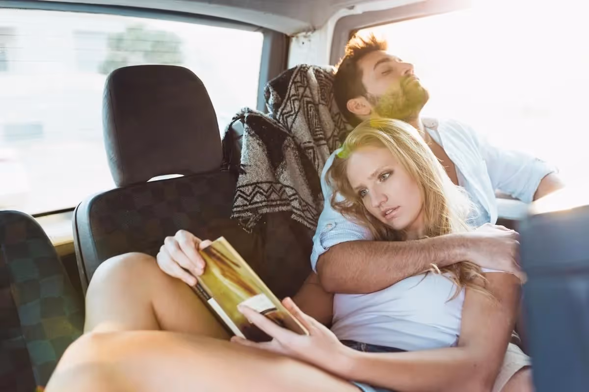 Un couple assis à l’arrière d’une voiture, la femme lisant un livre tandis que l’homme repose sa tête en arrière, profitant d’un moment de détente.