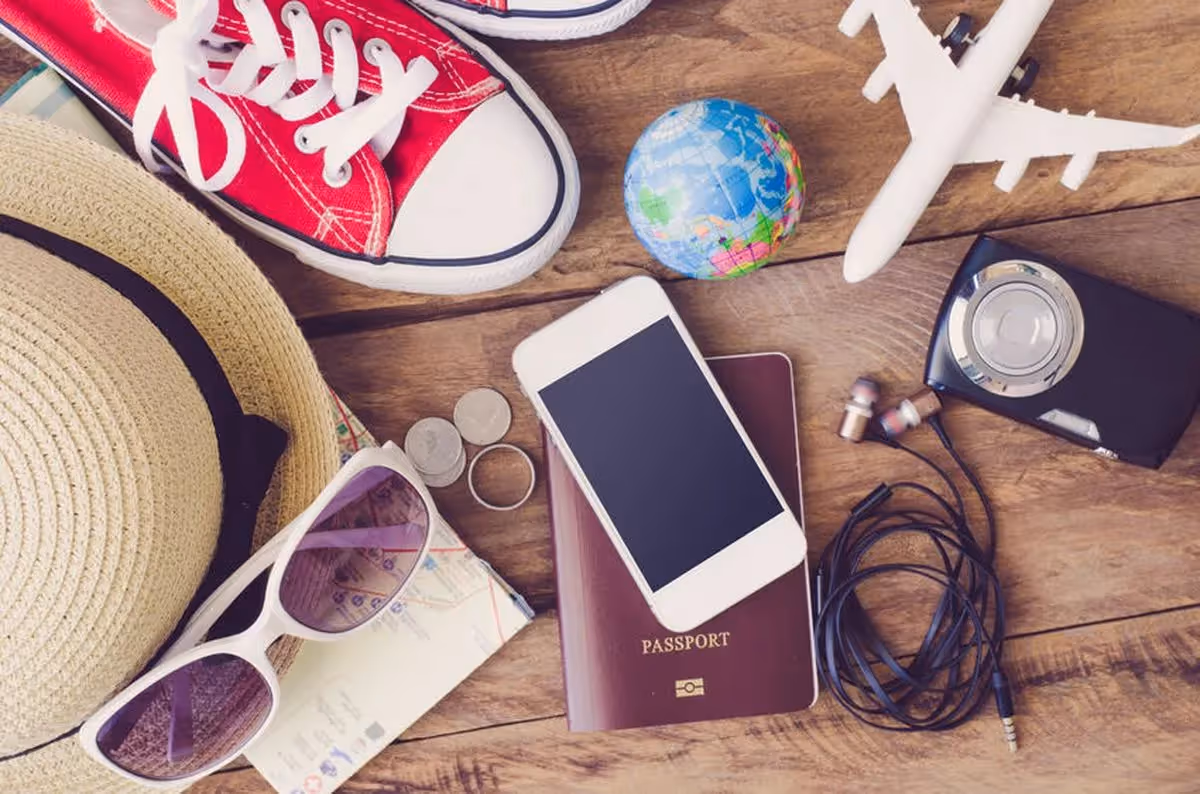 Accessoires de voyage posés sur une table : passeport, smartphone, appareil photo, écouteurs, globe, avion miniature, chapeau, lunettes de soleil, carte et chaussures rouges. 