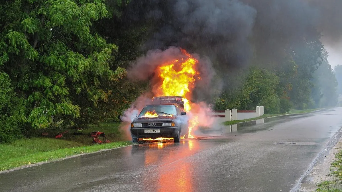 Voiture en feu sur une route mouillée, avec d’épaisses fumées noires et des flammes visibles à l’avant du véhicule