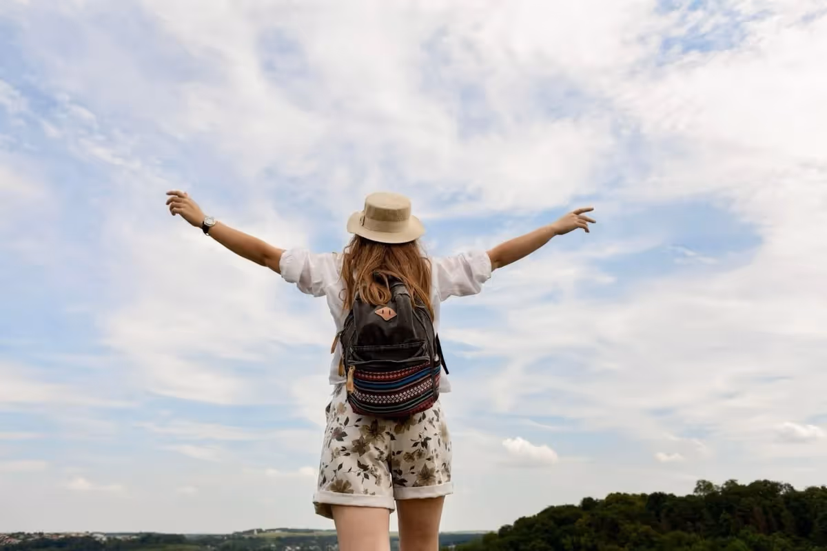 Voyageuse avec un chapeau et un sac à dos admirant la nature les bras ouverts, symbole de liberté et de voyage écoresponsable.