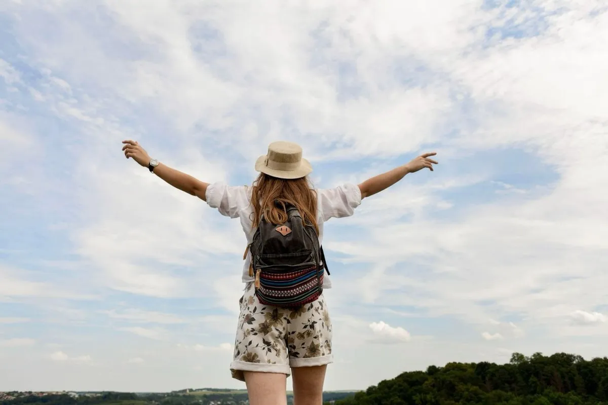 Voyageuse avec un chapeau et un sac à dos admirant la nature les bras ouverts, symbole de liberté et de voyage écoresponsable.