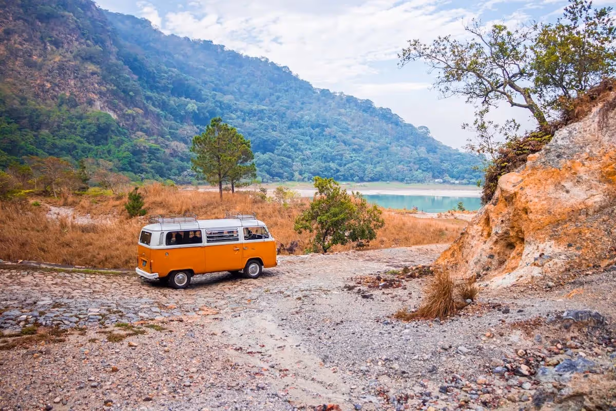Combi vintage orange garé sur un chemin de terre devant un lac de montagne entouré de forêt, en pleine nature.