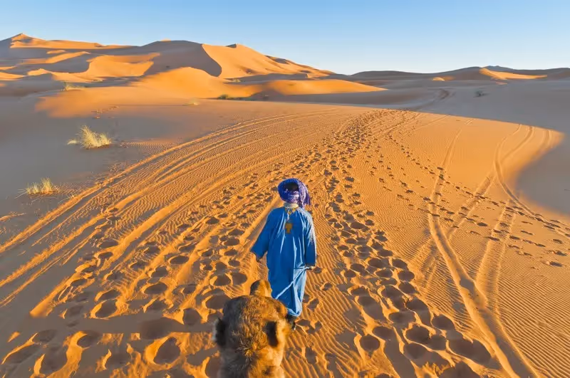 Homme en tenue bleue marchant dans les dunes dorées du désert de Merzouga, au Maroc.