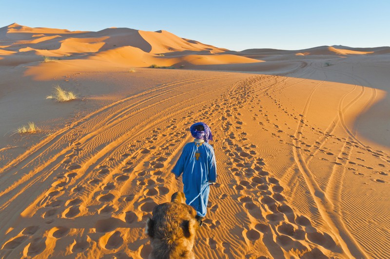 Homme en tenue bleue marchant dans les dunes dorées du désert de Merzouga, au Maroc.