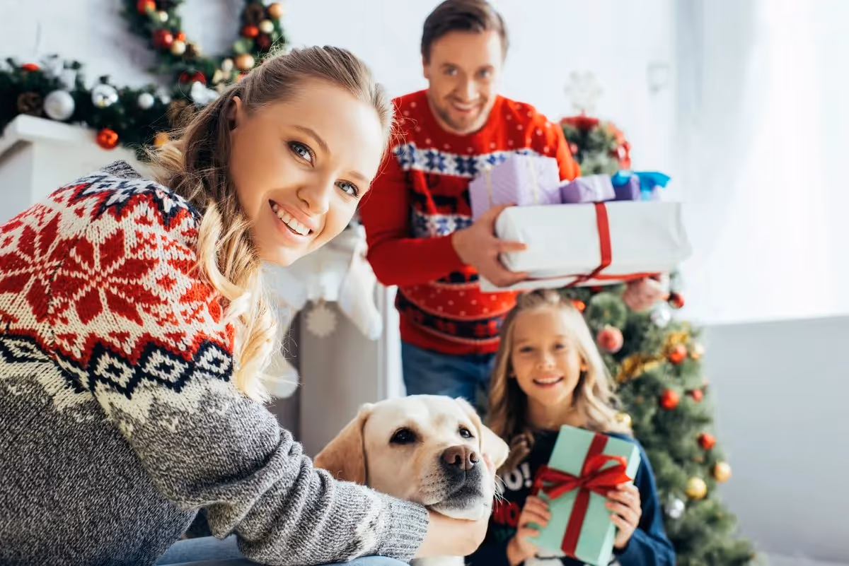 Famille souriante en pull de Noël offrant des cadeaux près du sapin avec leur chien, symbole de convivialité et d’esprit de fête.