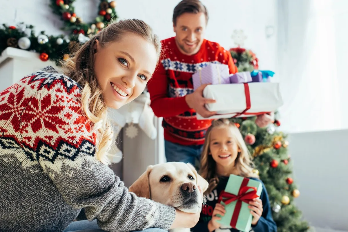 Famille souriante en pull de Noël offrant des cadeaux près du sapin avec leur chien, symbole de convivialité et d’esprit de fête.