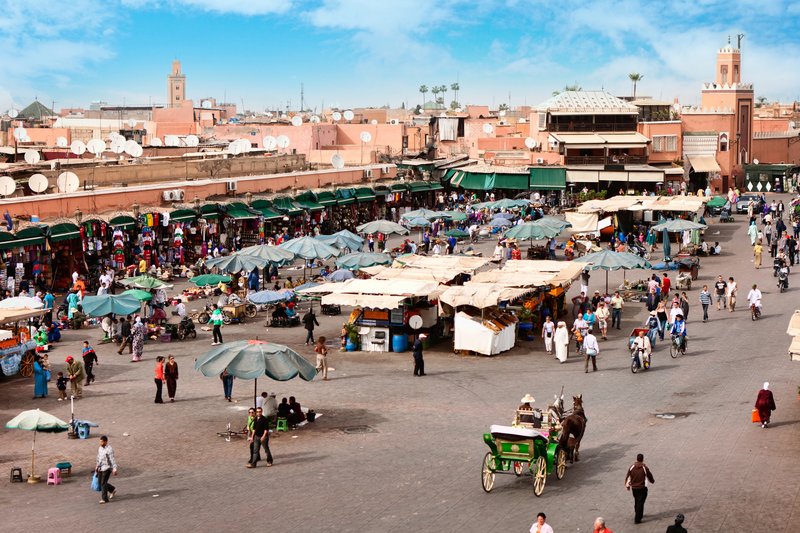 Vue animée de la Place Jemaa El-Fna à Marrakech, avec stands de marché, passants et ambiance locale en plein jour.