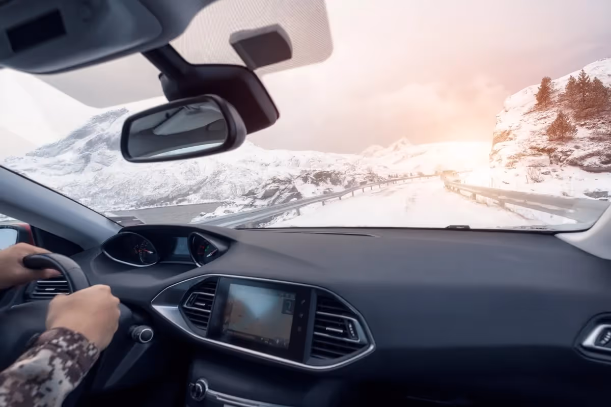 Vue depuis l’intérieur d’une voiture roulant sur une route enneigée en montagne, avec le conducteur au volant et un paysage hivernal au lever du soleil.