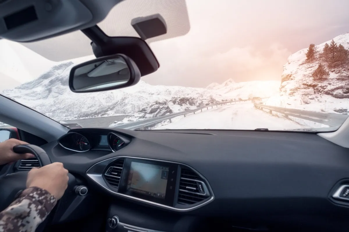 Vue depuis l’intérieur d’une voiture roulant sur une route enneigée en montagne, avec le conducteur au volant et un paysage hivernal au lever du soleil.