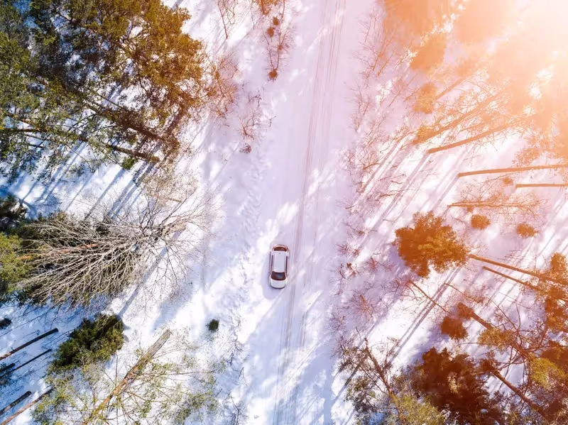 Vue aérienne d’une voiture roulant seule sur une route enneigée entourée de forêt.