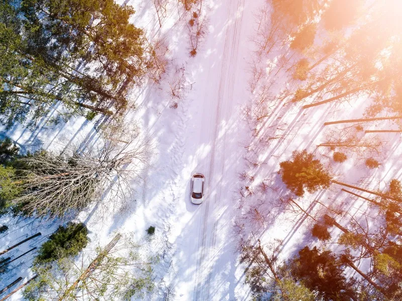 Vue aérienne d’une voiture roulant seule sur une route enneigée entourée de forêt.