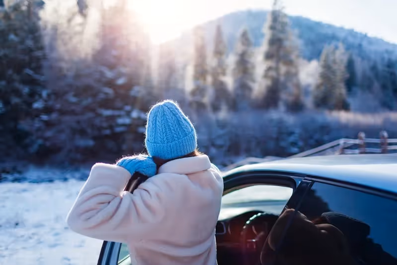 Personne en manteau et bonnet bleus sortant d’une voiture devant une forêt enneigée sous un soleil d’hiver.