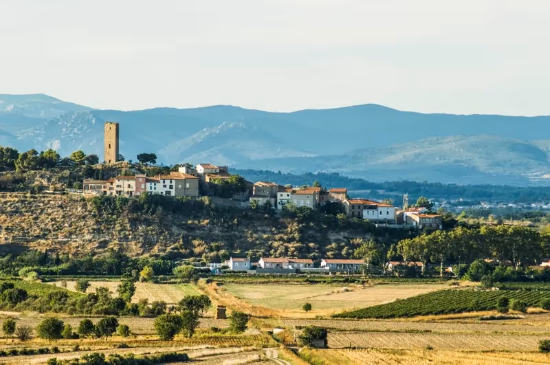 Vue d’un village perché en Occitanie, entouré de champs dorés et dominé par une ancienne tour, avec des montagnes bleutées en arrière-plan.