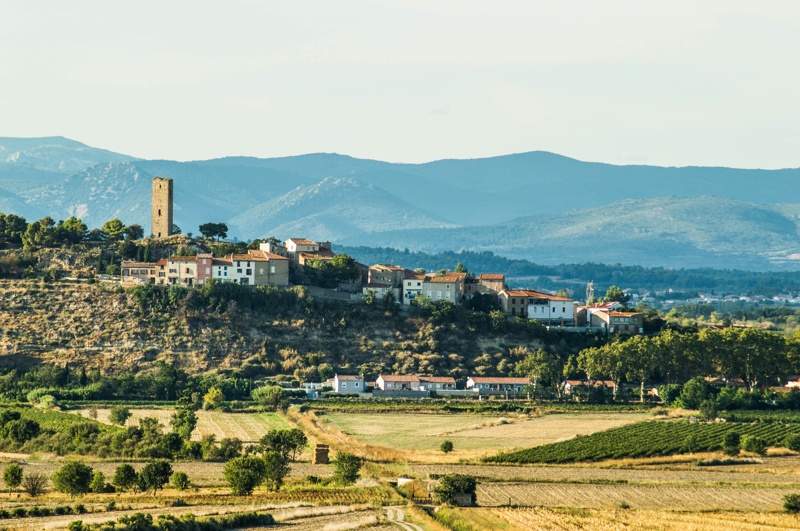Vue d’un village perché en Occitanie, entouré de champs dorés et dominé par une ancienne tour, avec des montagnes bleutées en arrière-plan.