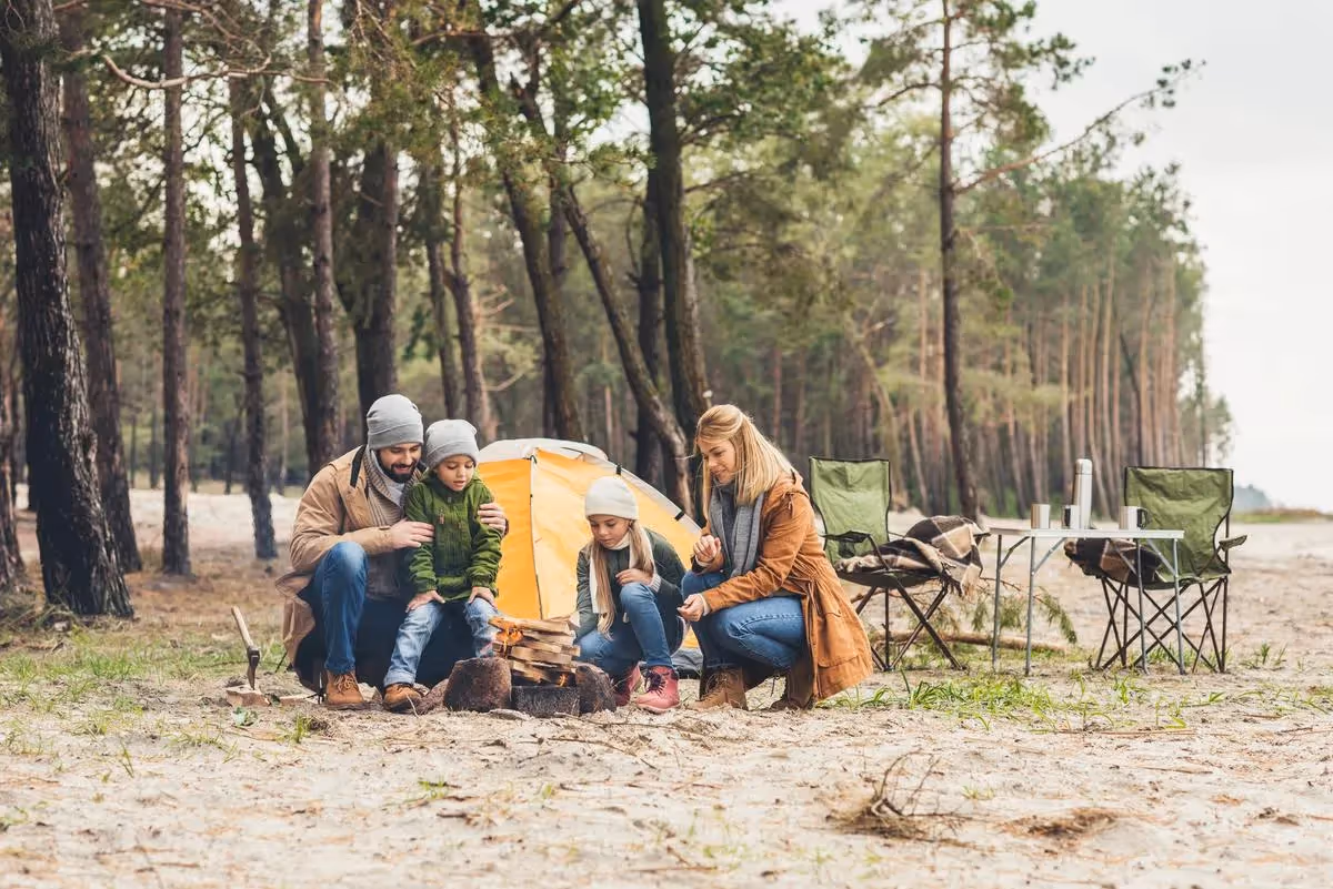 Famille en plein air installant un campement, avec deux enfants et leurs parents assis autour d’un petit feu devant une tente dans une forêt.