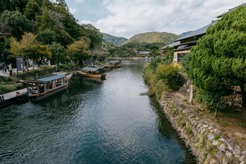 Bateaux traditionnels amarrés le long de la rivière à Arashiyama, entourés de collines verdoyantes et de maisons japonaises en bois.