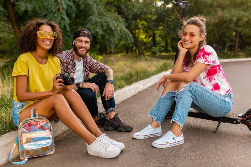 Trois amis souriants assis dans un parc avec un appareil photo et un sac à dos holographique, prêts pour un road trip