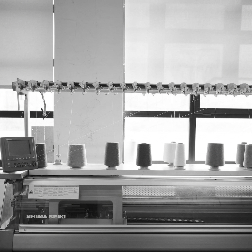 Black and white image of a Shima Seiki industrial knitting machine in a workshop with multiple large yarn cones arranged on top and threads feeding into the machine.