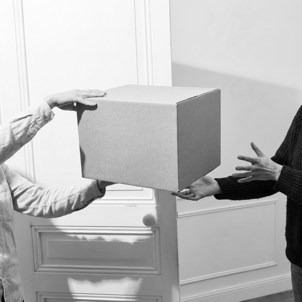 Two people exchanging a plain cardboard box indoors; one person wearing a light-colored shirt hands the box to another person wearing a dark sweater, with both sets of hands visible.
