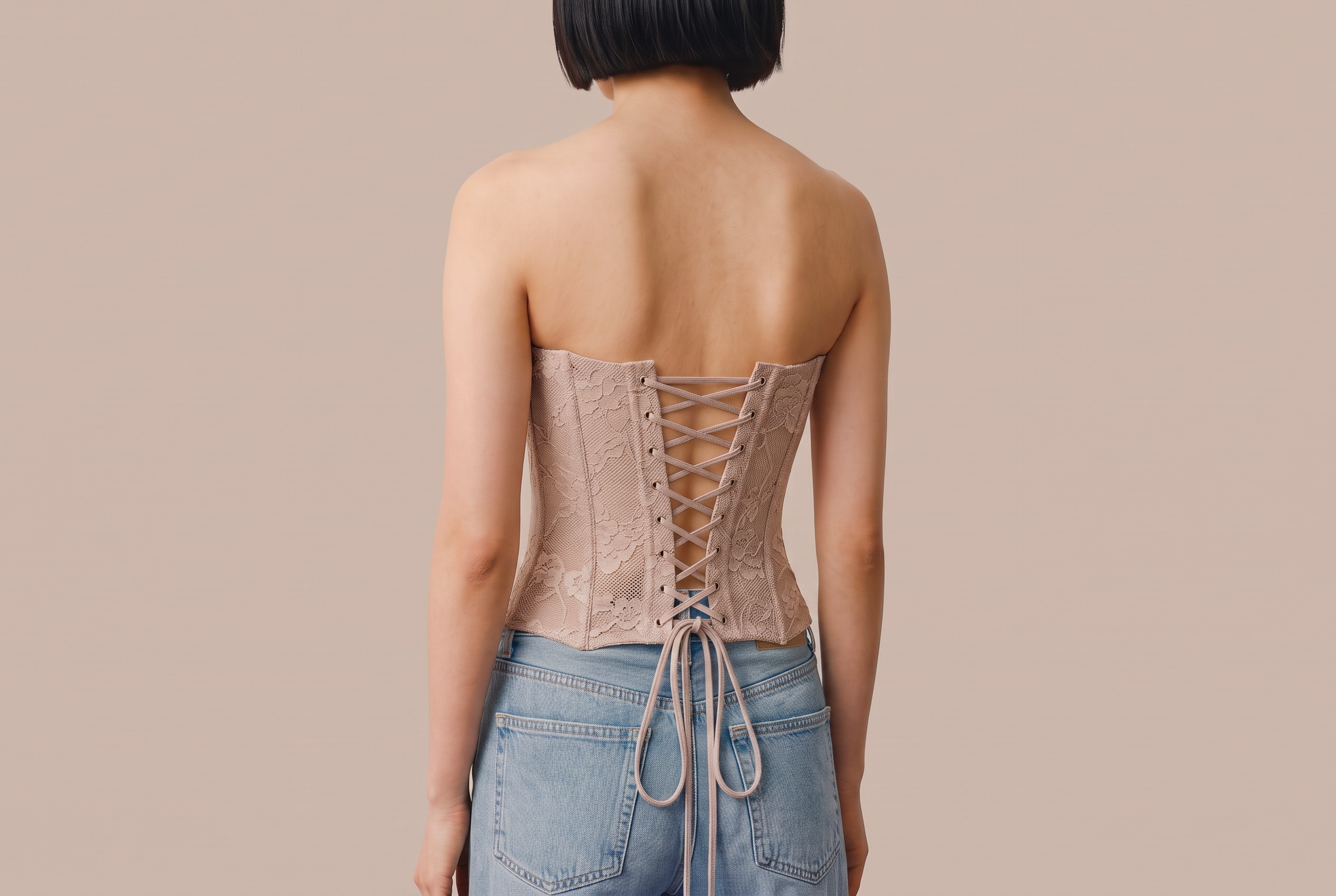 Rear view of a person wearing a light pink lace-up corset and light blue jeans against a plain beige background.