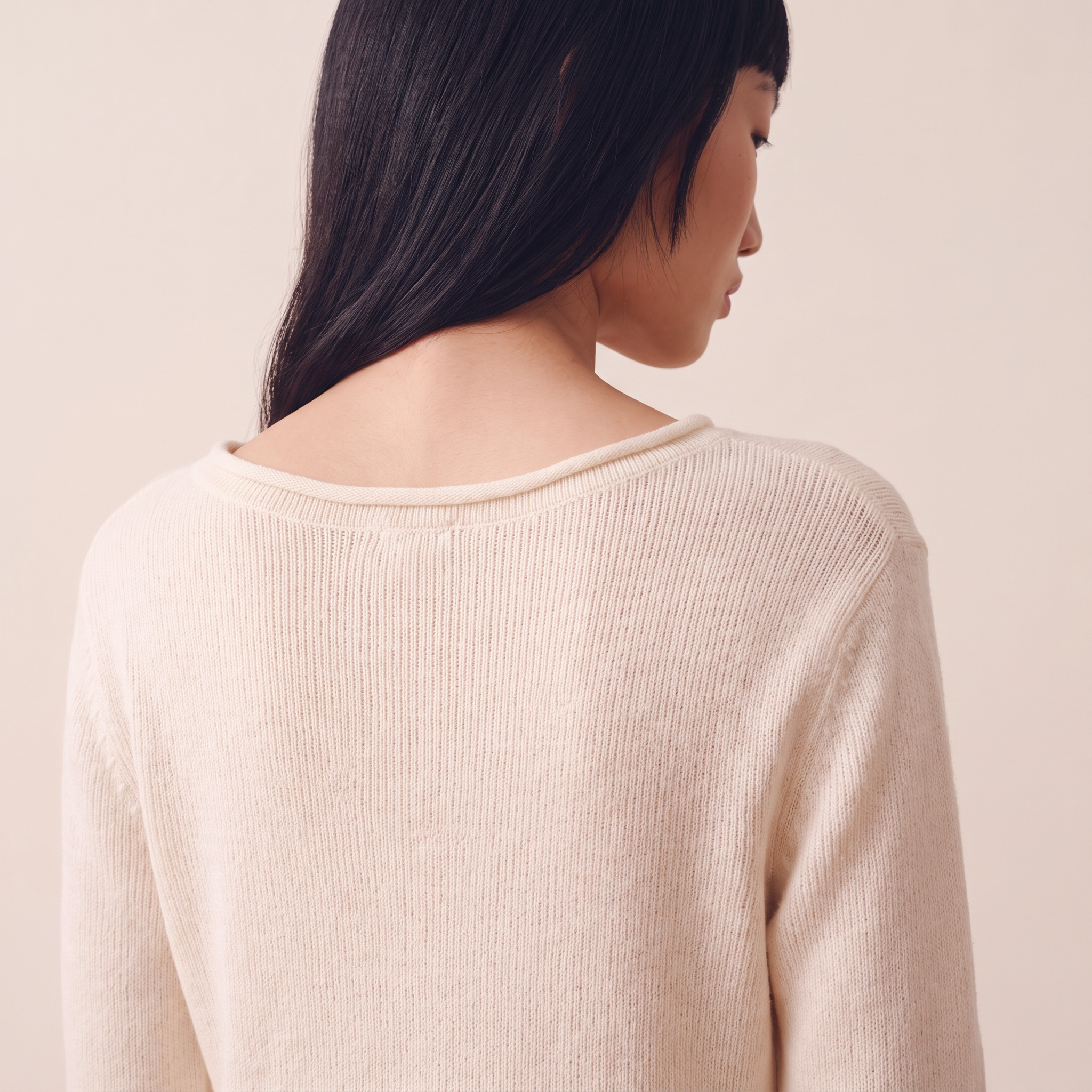Woman with long dark hair wearing a light beige knitted sweater viewed from behind against a pale background.
