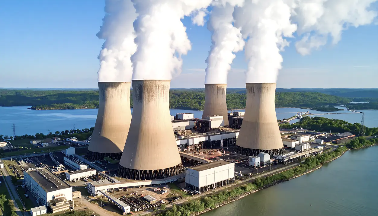 Aerial view of a power plant with four large cooling towers emitting thick white steam near a body of water and green hills.