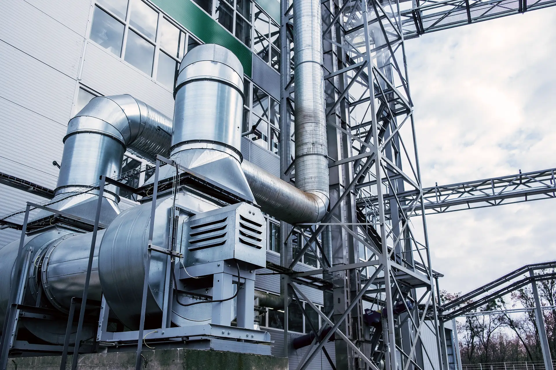 Large industrial metal ventilation and ducting system attached to a factory building under a cloudy sky.