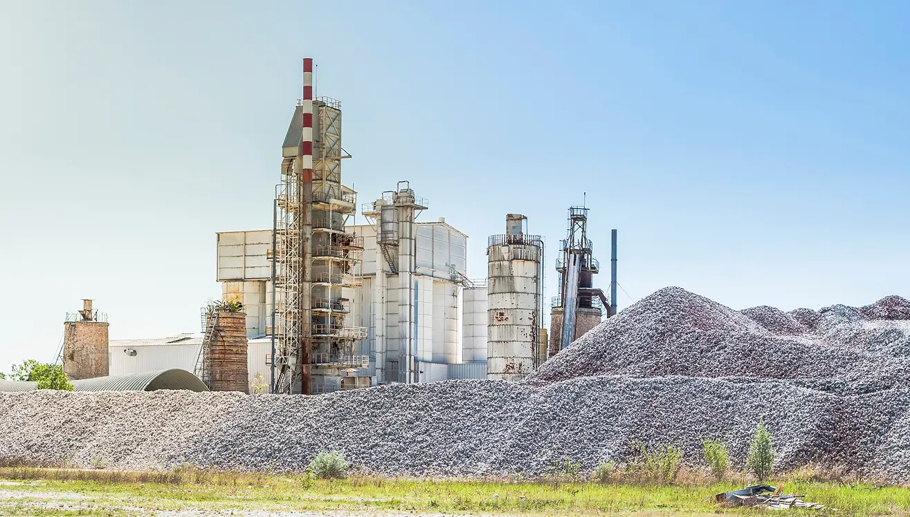 Industrial plant with large metal silos and chimneys behind expansive piles of raw material or mineral rocks under a clear blue sky.