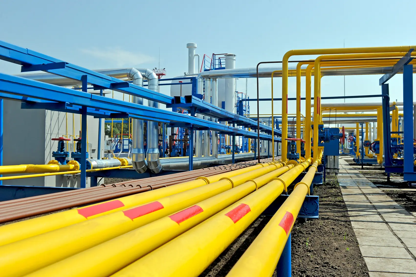 Industrial facility with parallel rows of yellow and blue pipelines and valves under a clear sky.
