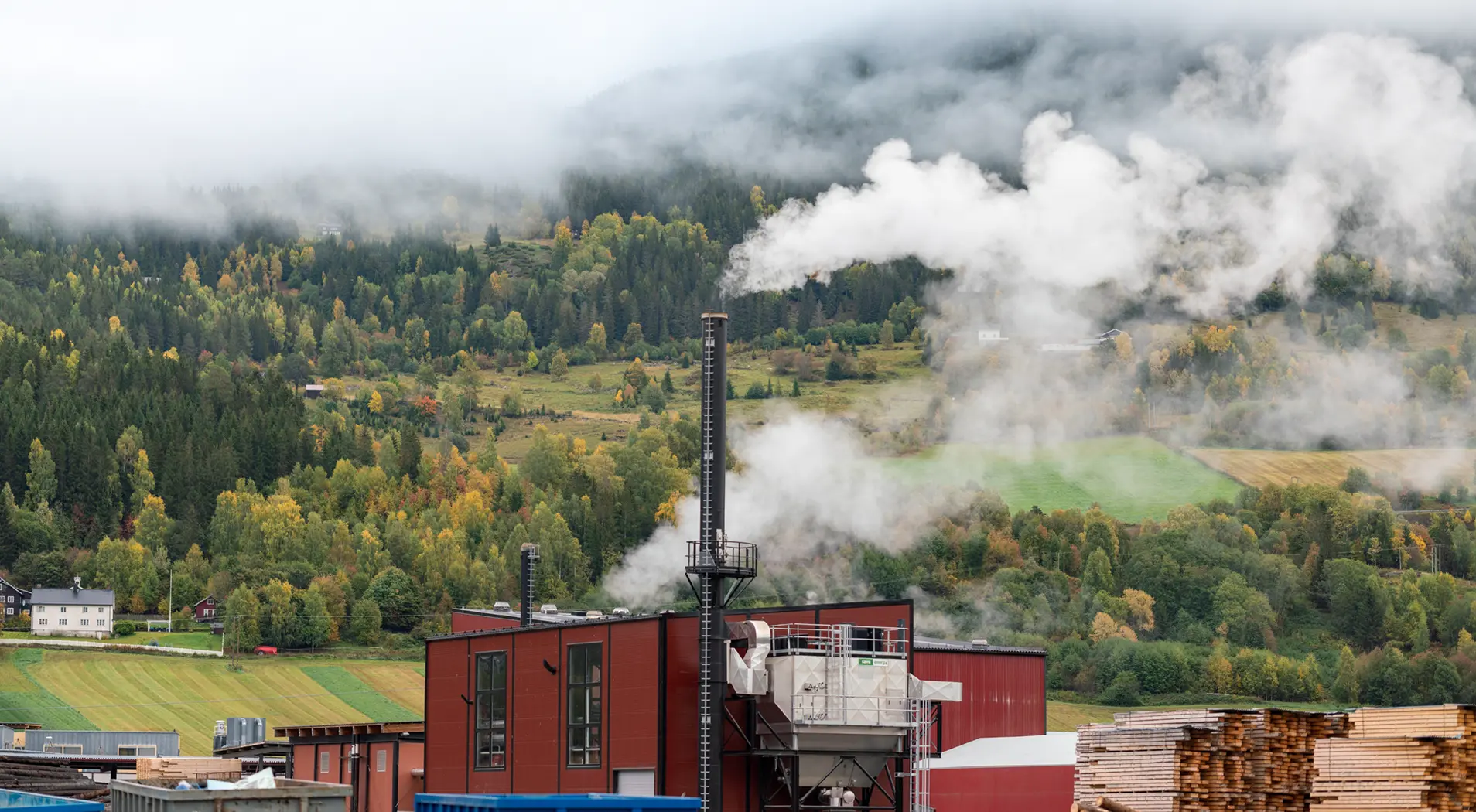 Industrial building with chimneys emitting white smoke in a green mountainous landscape with misty clouds.
