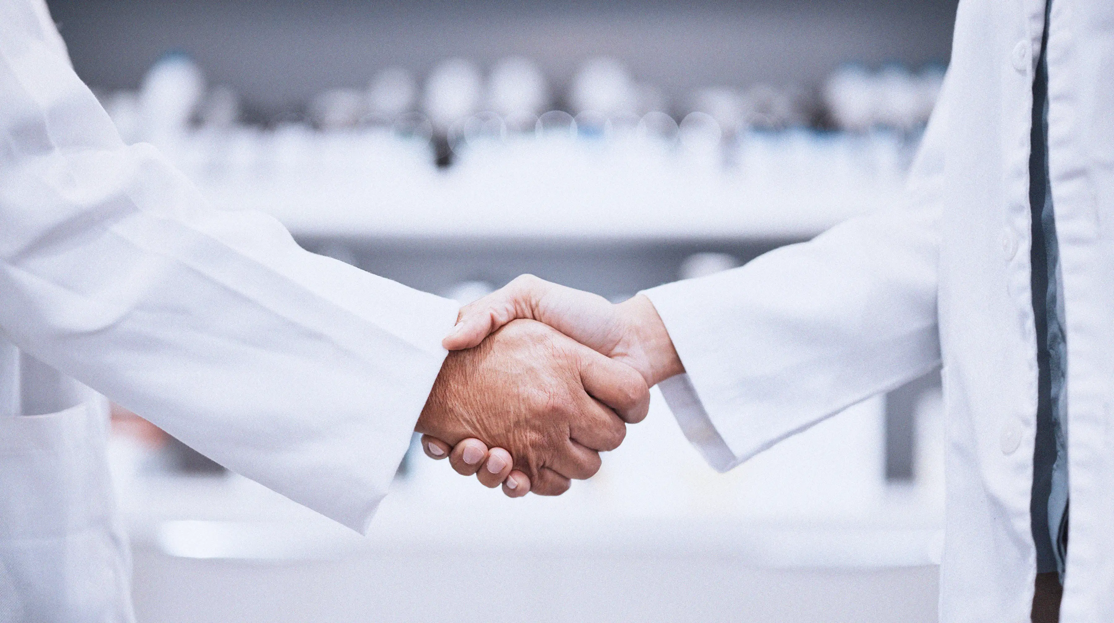 Two people wearing white lab coats shaking hands in a clinical or laboratory setting.