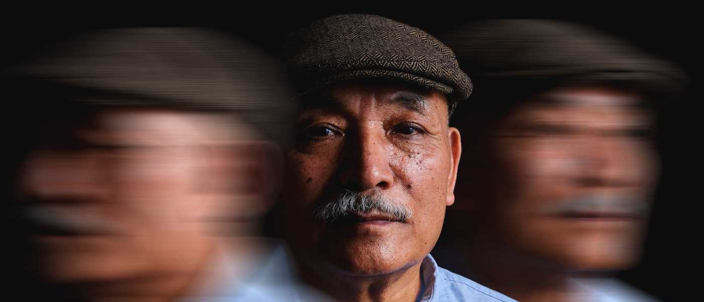 Portrait of an elderly man with a serious expression wearing a flat cap, showing visual motion blur on both sides to represent Parkinson's disease symptoms such as tremors or cognitive effects.