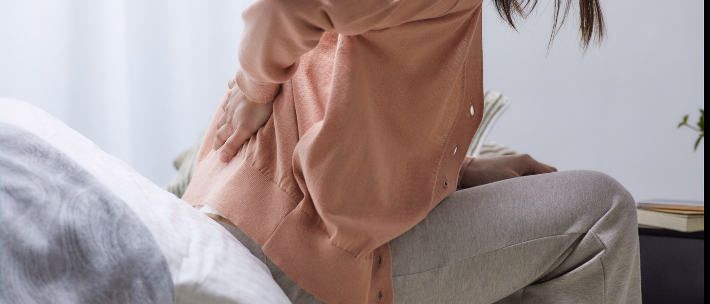 Woman sitting on bed holding lower back in pain, showing symptoms of lumbar degenerative disc disease (DDD)