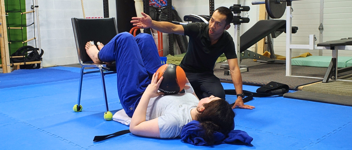 Physiotherapist guiding a patient through an LCL rehabilitation exercise on a clinic mat. The patient is lying on their back with knees bent and feet elevated on a chair, holding a medicine ball against their chest while performing a controlled bridge exercise. The therapist kneels beside them, providing instruction and monitoring form in a gym-based rehab setting.