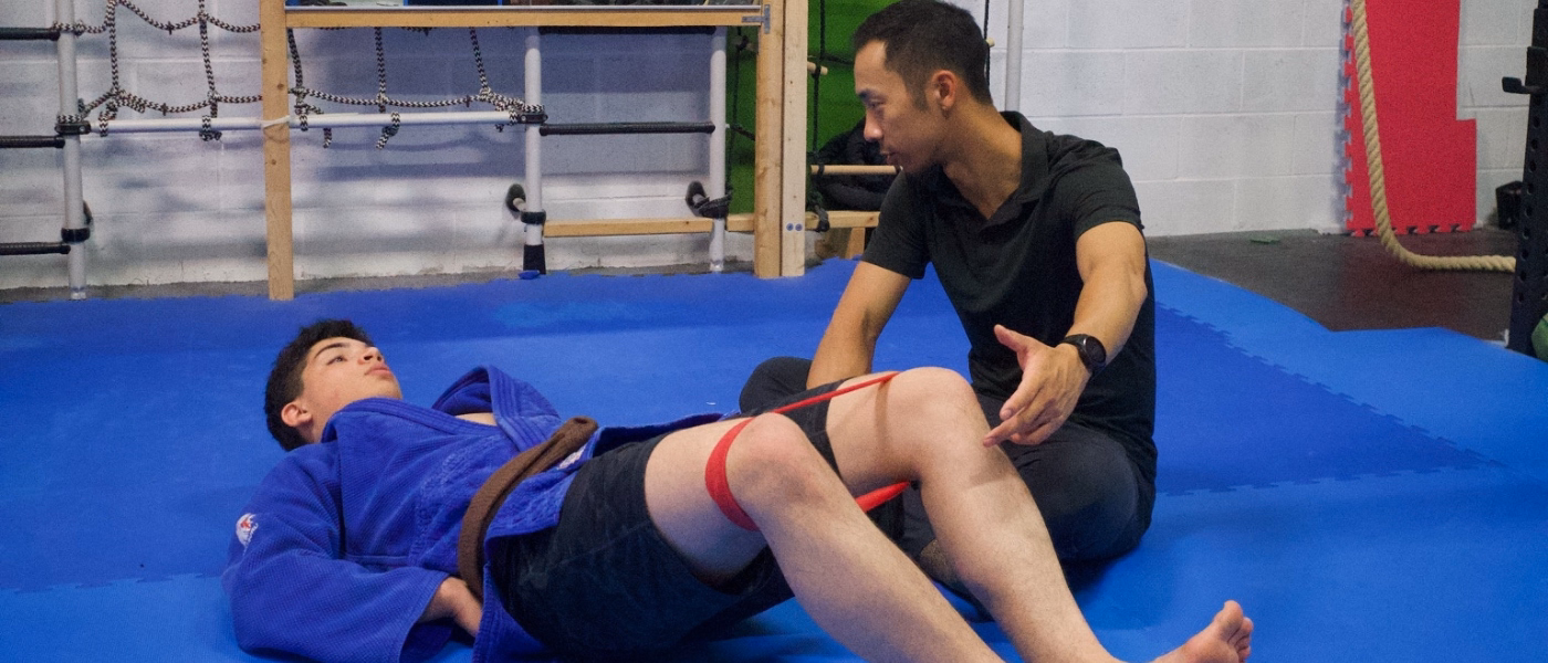 Physiotherapist instructing a patient performing a lower limb rehabilitation exercise for saphenous nerve entrapment. The patient is lying on their back on a clinic mat with a resistance band looped around their thighs, knees bent, while the therapist sits beside them providing guidance and monitoring leg alignment in a gym-based treatment setting.