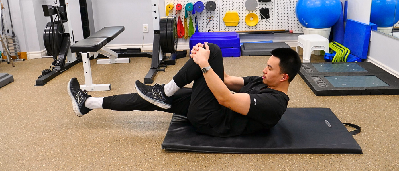 Physiotherapist demonstrating a controlled knee-to-chest exercise on a treatment mat as part of MCL sprain rehabilitation. The clinician lies on their back pulling one knee toward the chest while extending the opposite leg, emphasizing core engagement and lower limb control in a clinic gym setting.