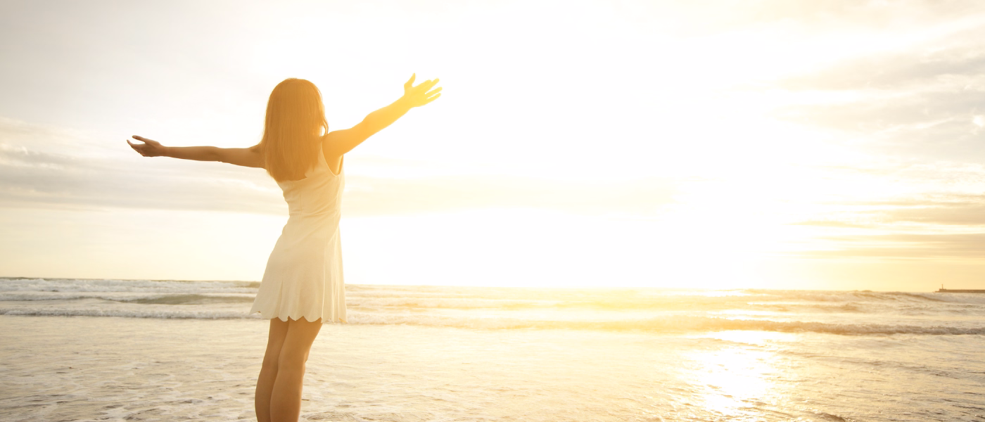 Woman standing on a beach at sunrise with arms open wide, symbolizing freedom, comfort, and confidence after pelvic organ prolapse recovery.