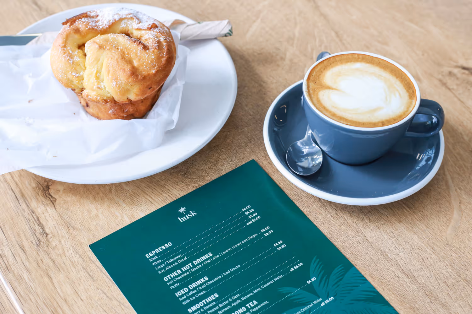 A green Husk Cafe menu on a wooden table, accompanied by a pastry on a plate and a cup of coffee
