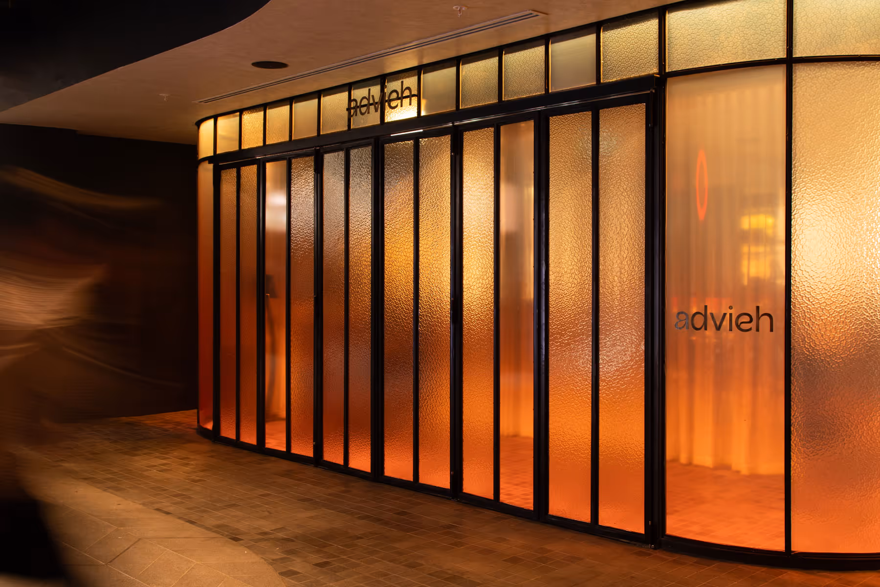 The illuminated, amber-toned glass storefront of Advieh restaurant with black metal framing.
