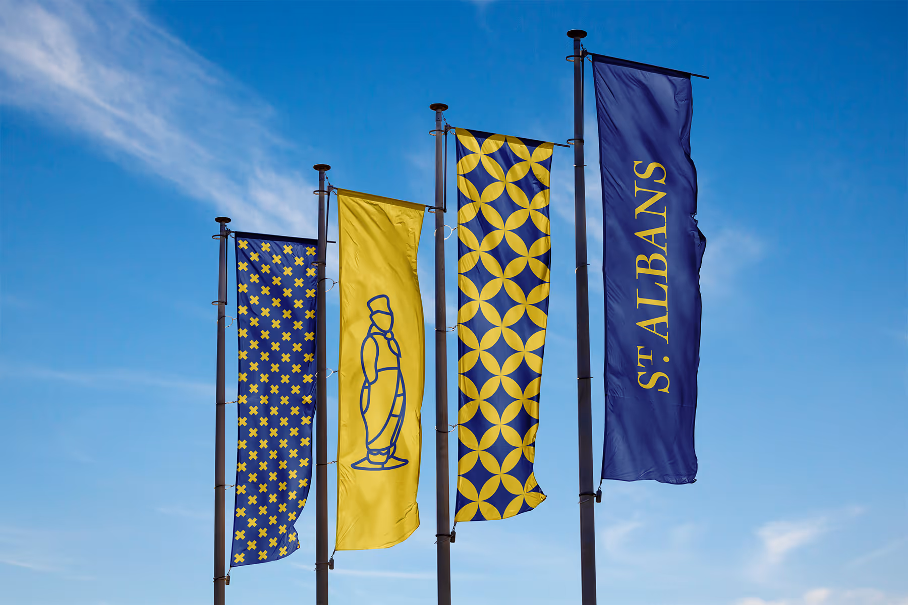 Four tall vertical flags against a blue sky; patterns include yellow crosses on blue, a blue outline of a statue on yellow, interlocking yellow circles on blue, and the text 'St Albans' in yellow on blue.