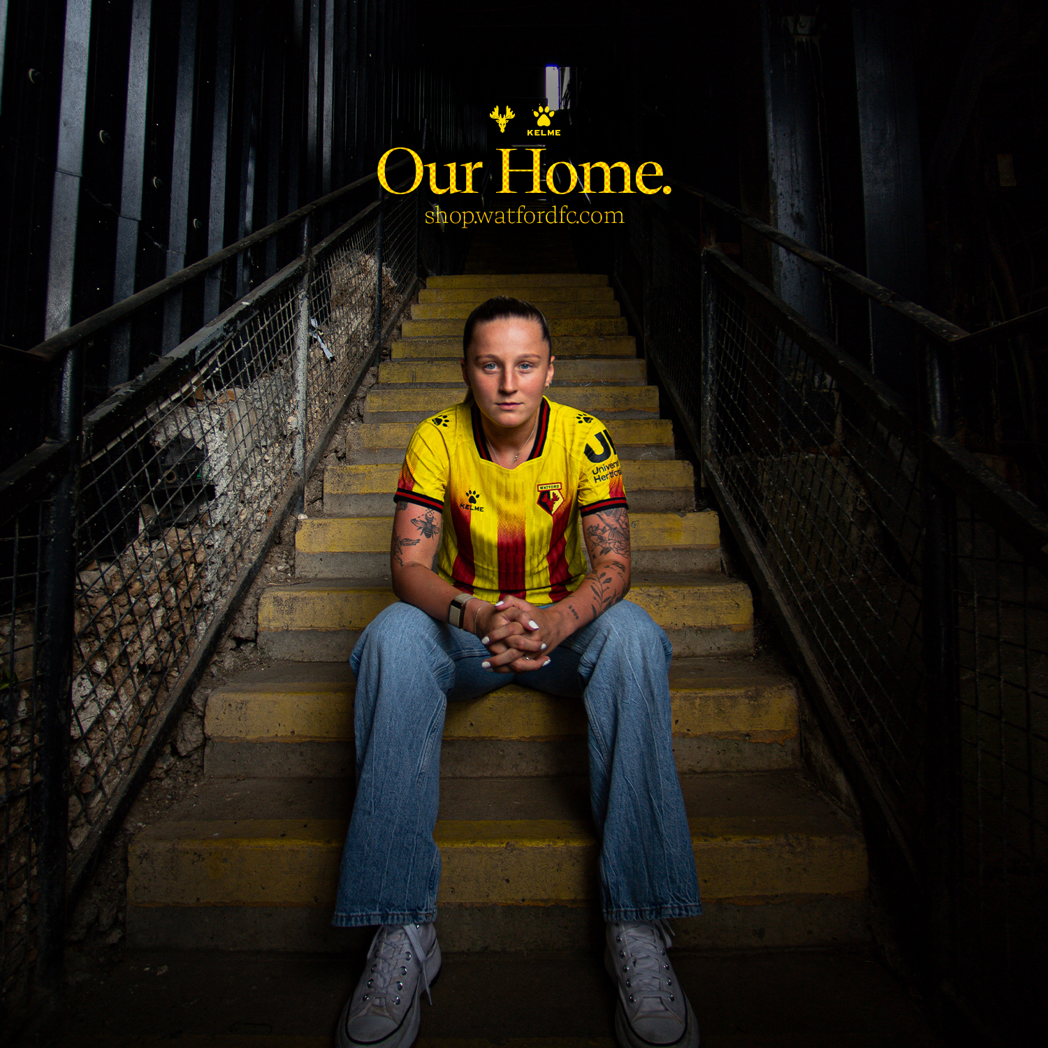 Football player, Anna Filbey, wearing a yellow and red Watford FC jersey sitting on concrete stairs in a dark, fenced corridor.