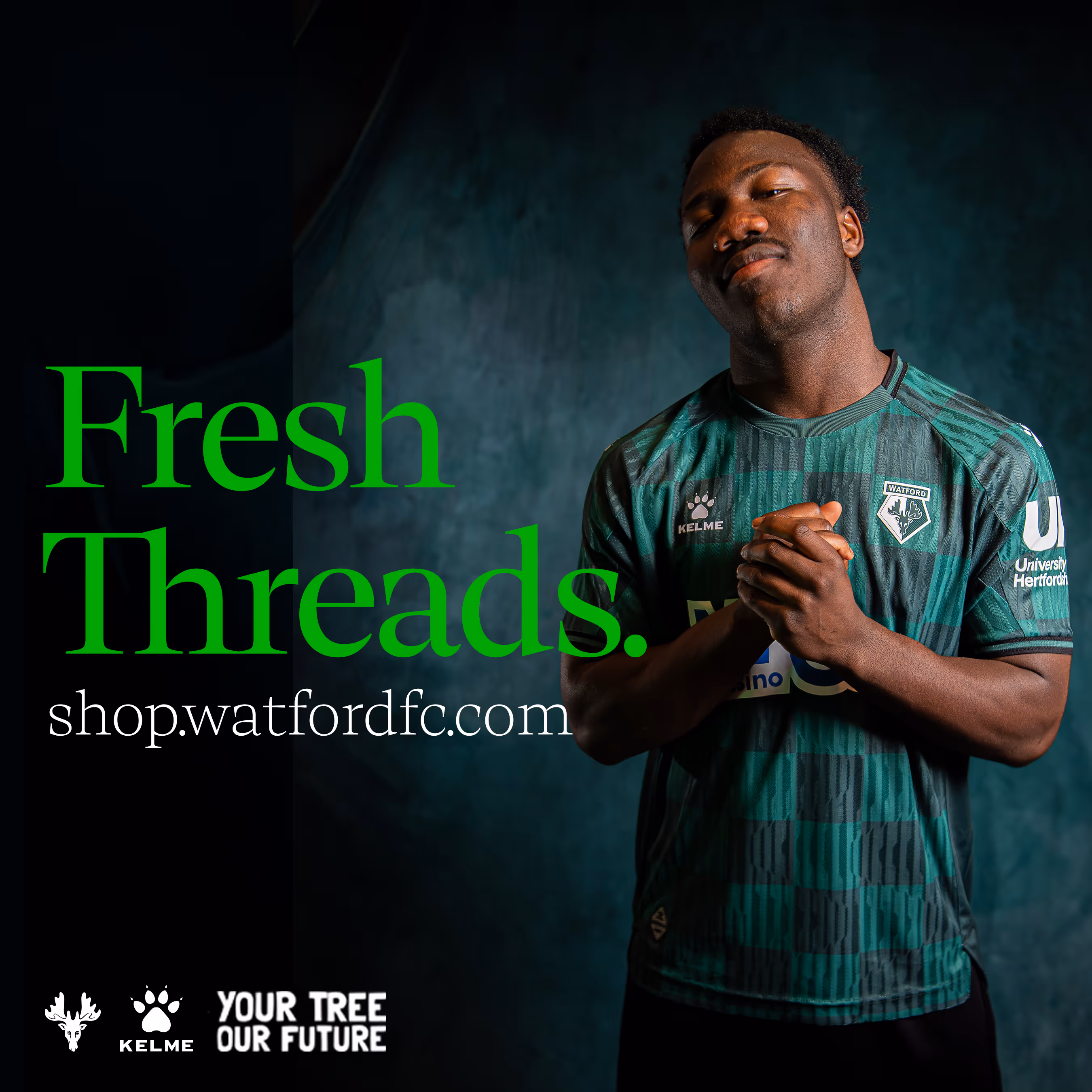 Football player, Nestory Irankunda, wearing green checkered Watford FC soccer jersey standing with clasped hands against a dark, textured background.