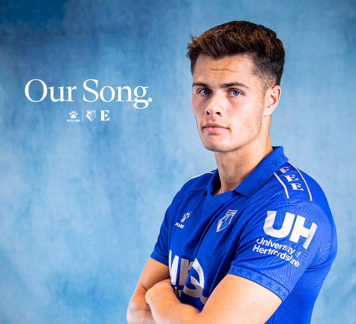 Football player, Luca Kjerrumgaard, in blue sports jersey, standing with arms crossed against blue background with text 'Our Song.'