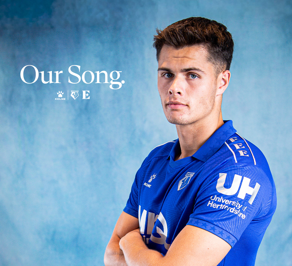 Football player, Luca Kjerrumgaard, in blue sports jersey, standing with arms crossed against blue background with text 'Our Song.'