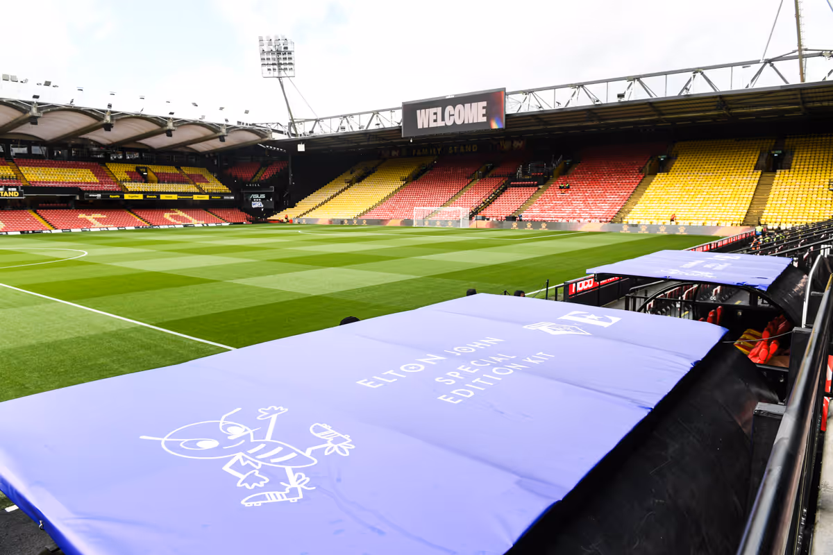 Empty football stadium, Vicarage Road, with yellow and red seats and a green pitch, featuring blue dugout covers labeled 'Elton John Special Edition Kit'.