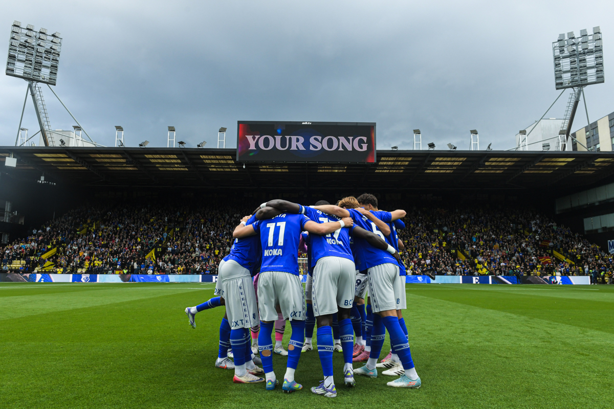 Watford team in blue jerseys huddling on the field inside a crowded Vicarage Road stadium under a sign that reads 'YOUR SONG'.